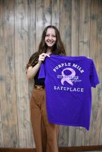 On the left, the front of the “Purple Mile Walk” T-shirts being sold now to benefit Safeplace can be seen in this photo of Safeplace Director Sandra Ells. Right, Safeplace Victim Advocate Avery Williams holds up one of the shirts showing the back screenprint. Please see the ad on this page and order your T-shirt now to wear at the event.